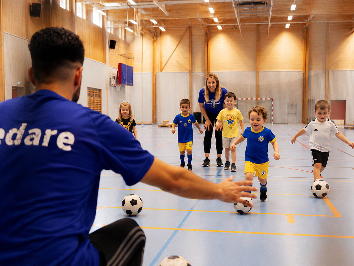 En grupp små barn övar fotboll i en idrottshall med ledare