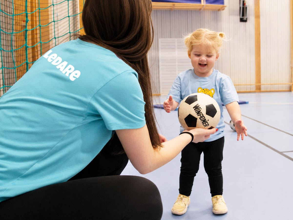 Ledare håller fram fotboll till litet leende barn.
