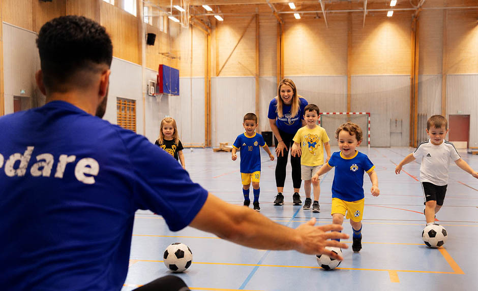 En grupp små barn övar fotboll i en idrottshall med ledare